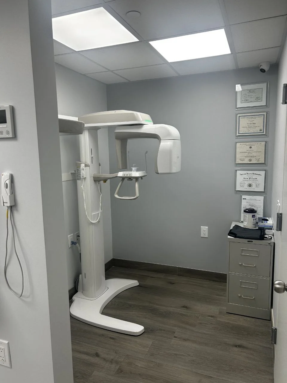 Imaging room at BP Smiles Orthodontics with a panoramic 3D cone-beam X-ray unit and Dr. Pinhasov's framed credentials on the wall.